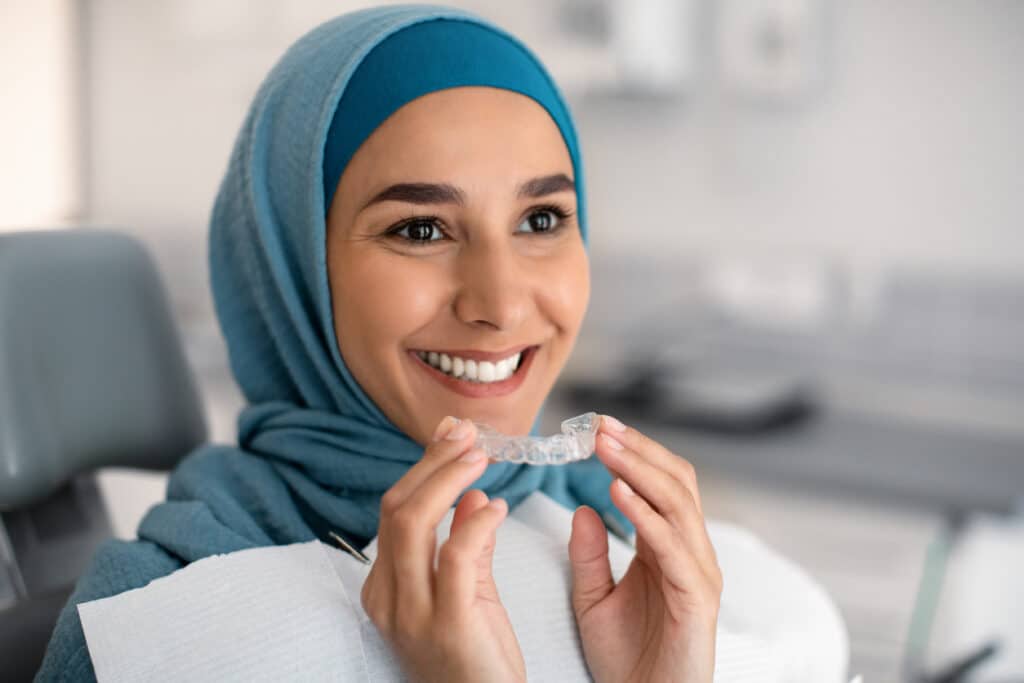 Orthodontics Concept. Smiling Muslim Woman In Hijab Holding Invisalign Or Invisible Braces While Sitting At Dentists Chair In Clinic, Islamic Female Using Clear Dental Aligner For Teeth Correction